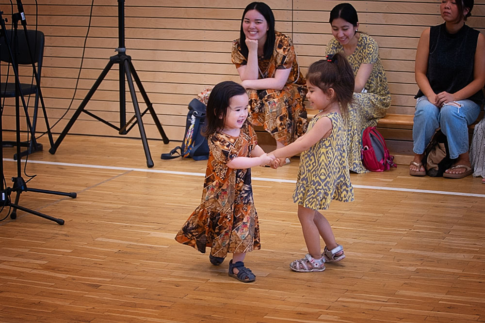 Zwei kleine Mädchen in Kleidern tanzen mit angefassten Händen im Kreis, dahinter ihre lächelnden Mütter in denselben Kleidern.