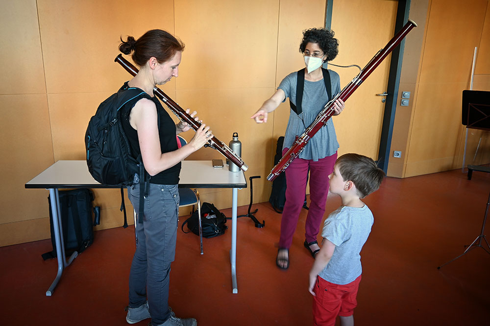 Eine Lehrerin mit Fagott lässt eine Frau dieses Instrument ausprobieren. Ihr kleiner Sohn steht staunend daneben.