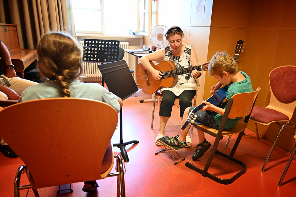 Blick in einen Raum mit zwei Kindern mit Konzertgitarren auf dem Schoß, einer versucht zu spielen. In der Mitte sitzt die Lehrerin, ebenfalls mit einer Gitarre.