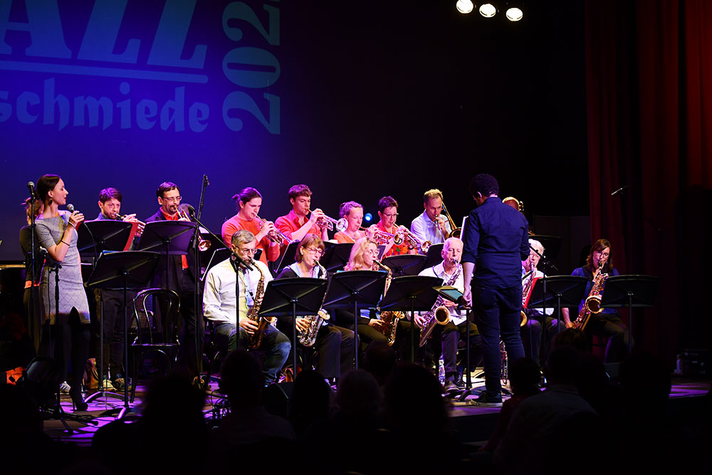 Die Bixband spielt auf der Bühne, links die Sängerin Franzi. Davor steht der Leiter der Band Philip Sindy. An der Bühnenrückwand liest man JazzSchmiede 2022.