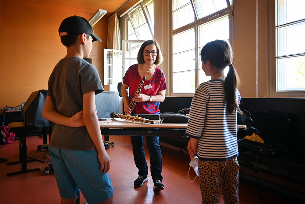 An einem Tisch stehen ein Junge und ein Mädchen und lauschen der Lehrerin, die ihnen etwas auf der Oboe vorspielt.