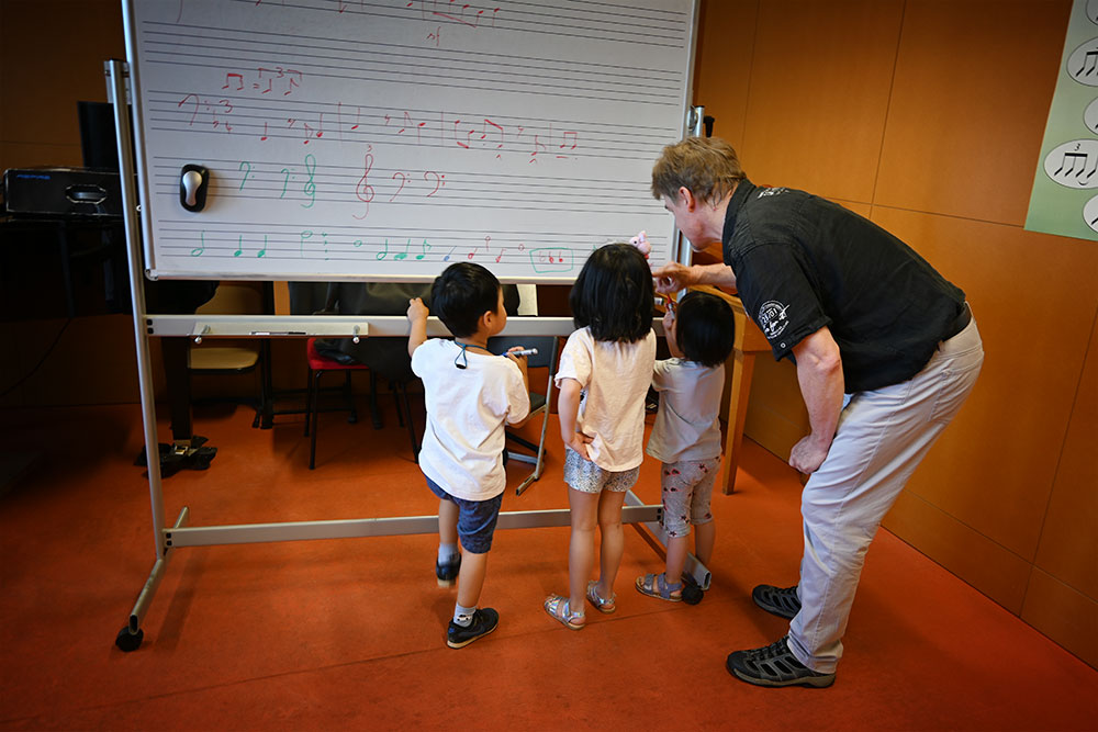 An einem White Board mit Notenlinien und Noten stehen drei kleine Kinder. Ein Lehrer zeigt ihnen etwas.
