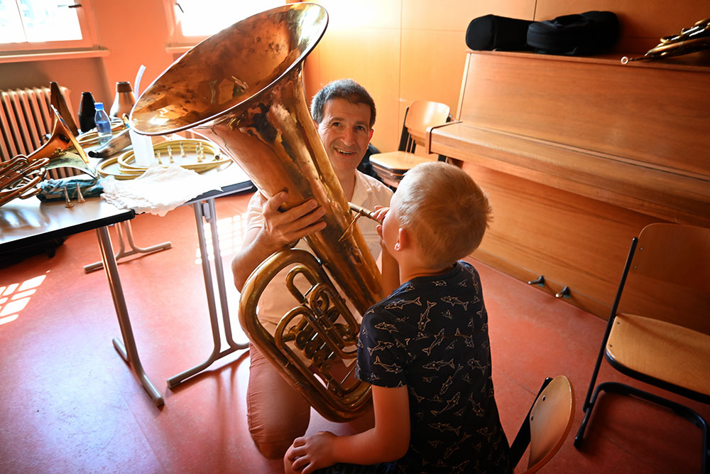 Ein Junge bläst im Vordergrund in eine große Tuba, die der Lehrer ihm hält.