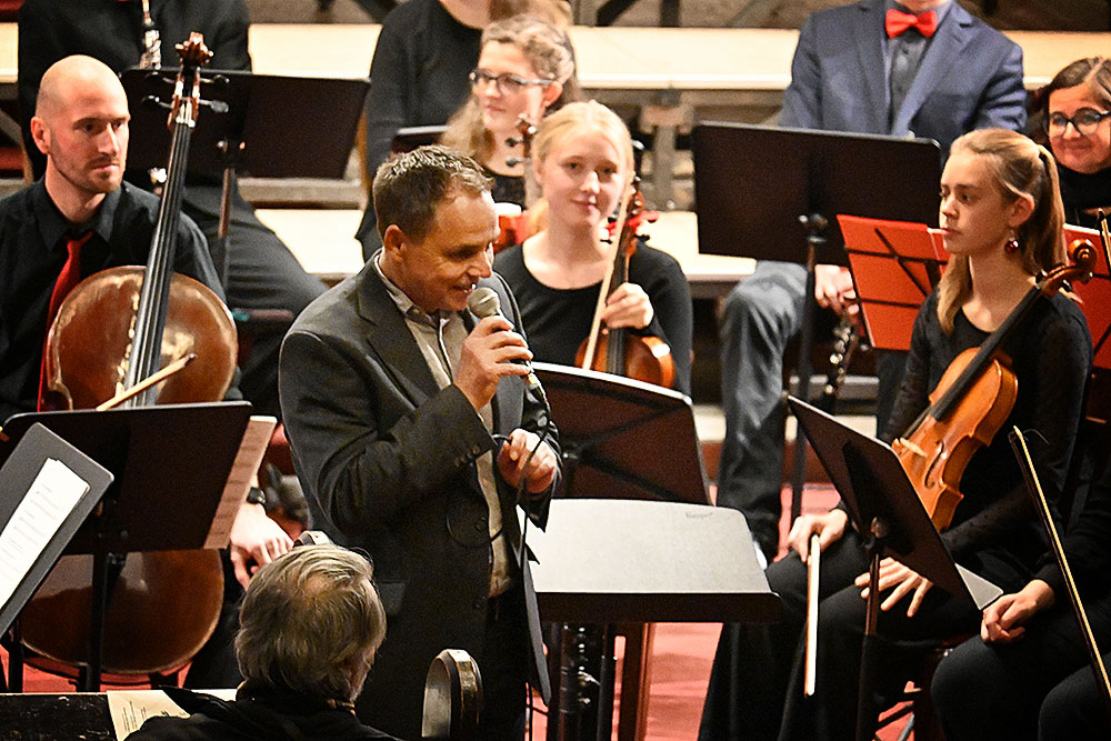 Musikschulleiter Olaf Hengst mit Mikrofon vor den Musiker:innen des Sinfonieorchesters