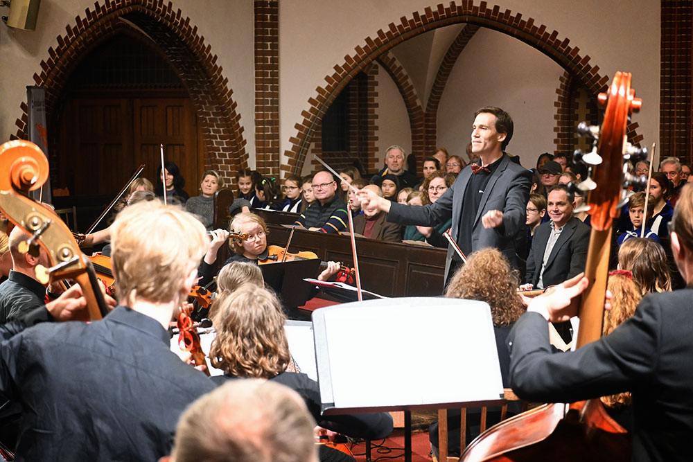 Foto von der Bühne über das Orchester ins Publikum. Man sieht Till Schwabenbauer mit einem Lächeln dirigieren.
