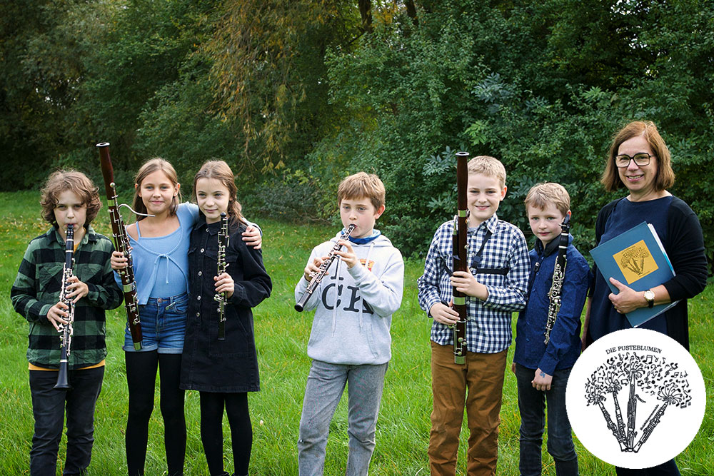 Sechs Kinder mit Klarinetten, Fagotte und Oboen stehen auf einer Wiese in einer Reihe, ihre Lehrerin rechts. Dahinter Bäume und Büsche eines Parks.