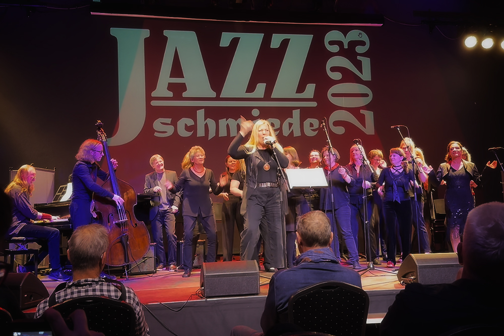 BerlinJazzSingers in Aktion auf der Bühne, im Hintergrund die Aufschrift "JazzSchmiede 2023