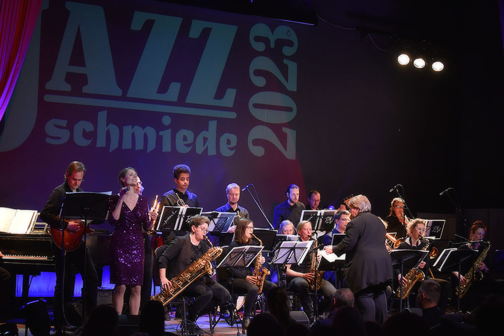 Gesamtansicht der BigBand Just Mad. Im Vordergrund die Leiterin der Band und die Sängerin. Im Hintergrund sieht man den Schriftzug JazzSchmiede 2023.
