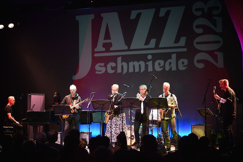 Von links nach rechts auf der Bühne: Pianist, Gitarrist, Saxophonistin, Trompeterin, Saxophonist, Bassist. Im Hintergrund das große Logo der JazzSchmiede 2023