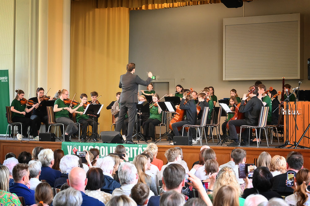 Das Orchester sitzt auf der Bühne und spielt, der Dirigent davor. Im Vordergrund sieht man einen Teil des Publikums.