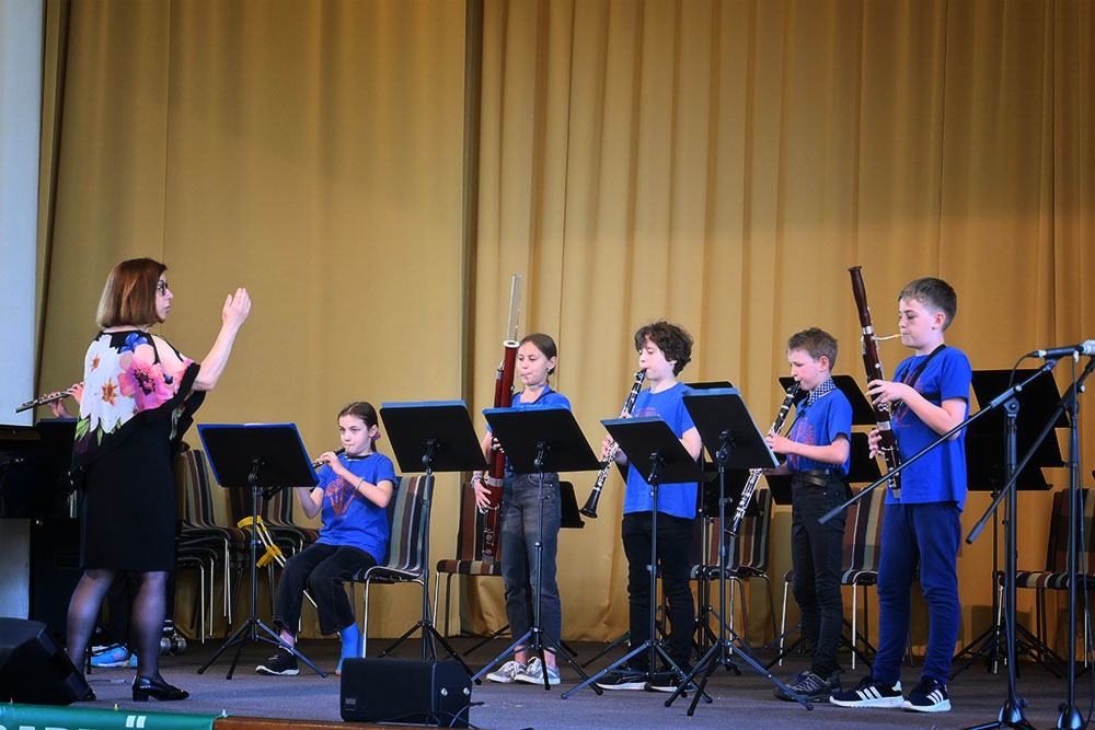 Fünf kleine Musikanten in blauen T-Shirts spielen Flöte, Klarinetten und Fagotte auf der Bühne vor einem beigen Vorhang, dirigiert von ihrer Lehrerin, die links im Bild steht.