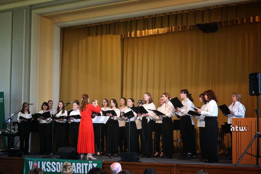 Der russische Chor - etwa 20 Frauen - singt auf der Bühne, schwarze Notenmappen in den Händen, weiße Blusen und schwarze Röcke/Hosen an. Vor ihnen dirigiert die Chorleiterin im roten Kleid.