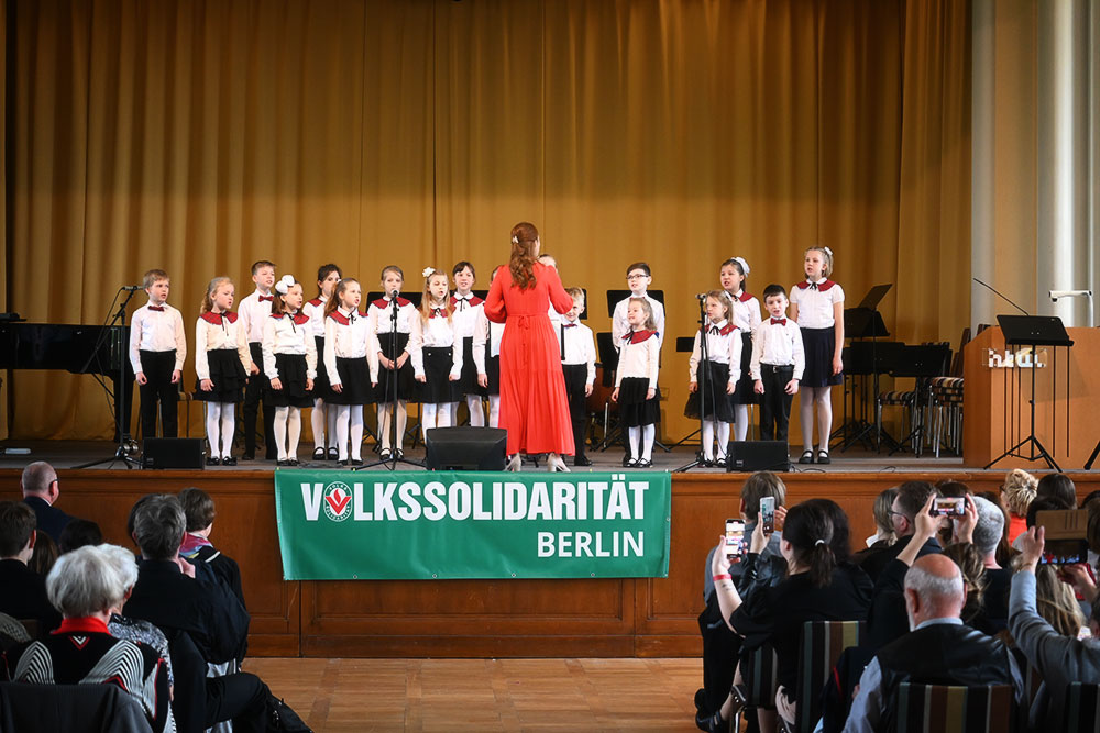 Die 18 Kinder des russischen Chores mit weißen Blusen mit rotem Kragen und schwarzen Röcken/Hosen singen auf der Bühne, dirigiert von ihrer Chorleiterin im roten Kleid. Davor das Publikum.