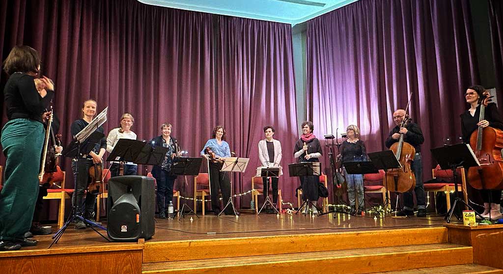 Das Ensemble Rock Barock steht mit ihren Instrumenten im Halbkreis auf einer Bühne, im Vordergrund eine Holztreppe, im Hintergrund lila Vorhänge. Links steht die Ensembleleiterin Sigrun Pleißner.