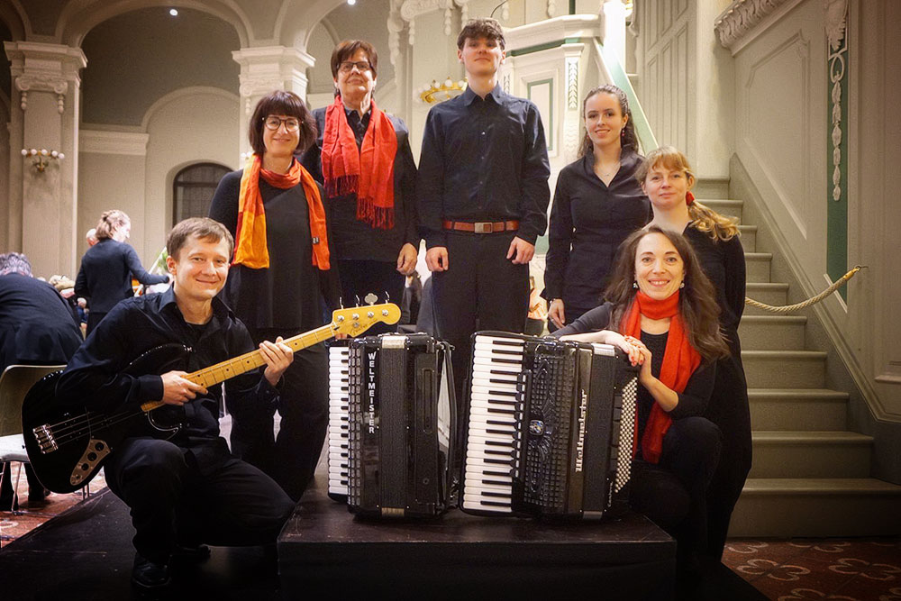 Sieben Mitglieder des Akkordeonensembles stehen hinter zwei schwarzen Akkordeons, links kniet der Bassgitarrist.
